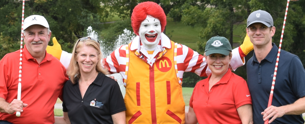 Ronald mcdonald standing on golf course posing with man and woman on left side, and another man and woman on right side. Both men are holding a golf club.